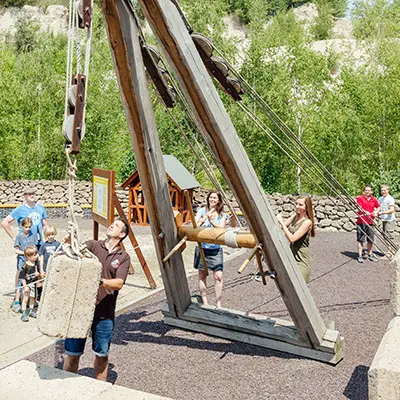 Besucher heben einen Steinblock mithilfe eines römischen Holzkrans in der Antiken Technikwelt des Römerbergwerk Meurin